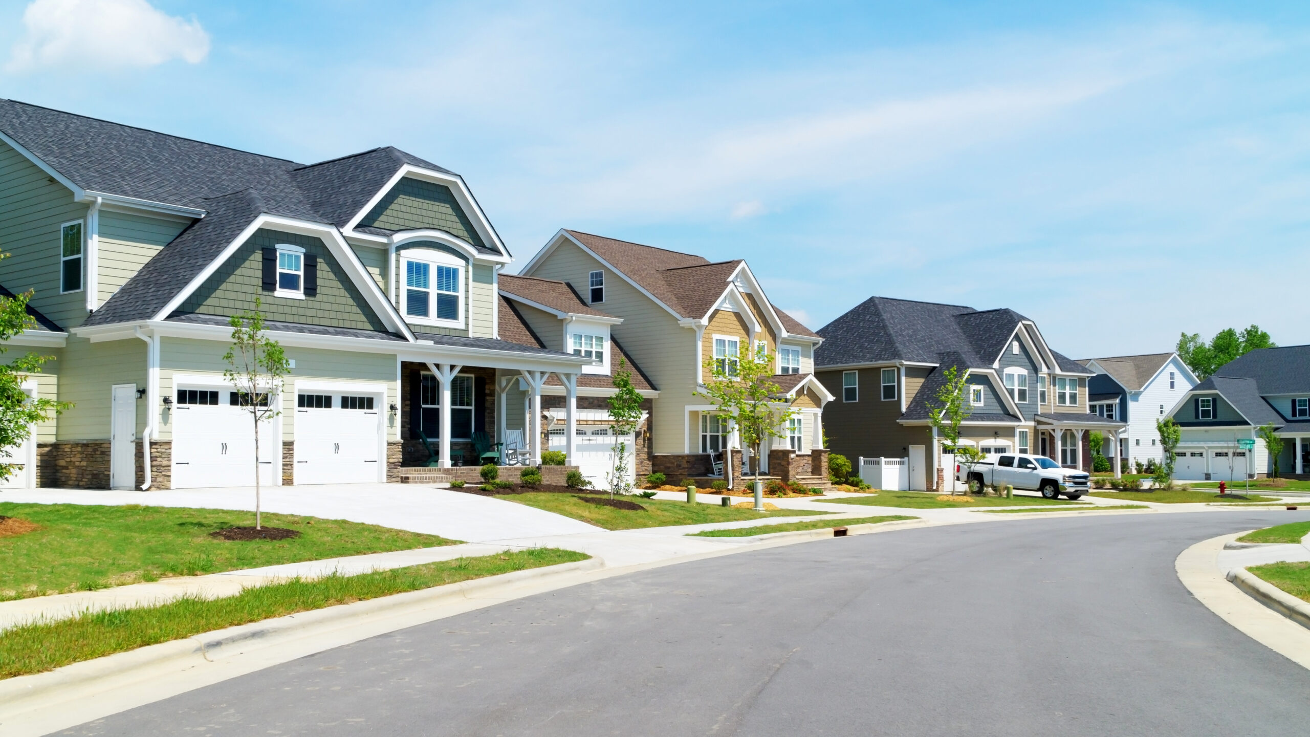 A suburban street with newly built, two-story houses with garages and well-maintained lawns. The houses are painted in various neutral colors and have modern architecture. The street is clean and curved, with young trees planted along the sidewalks.