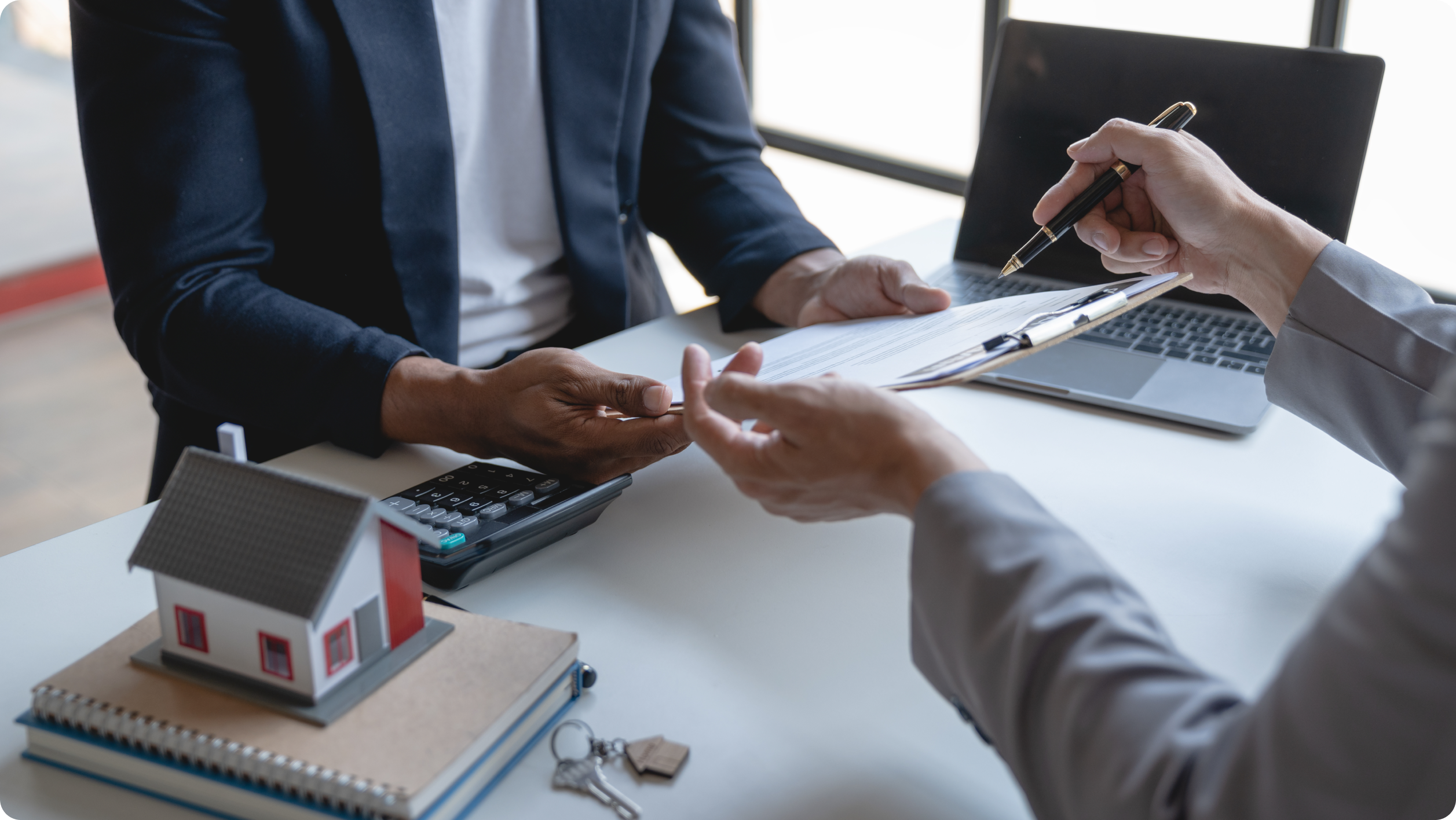 Two people at a table laden with paperwork, a laptop, and a small house model discuss opening an escrow account. One person points to documents on a clipboard while a calculator lies nearby, suggesting negotiations about real estate or financial transactions.