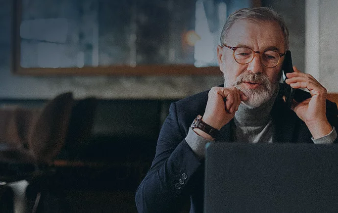 A bearded man with glasses, wearing a dark suit and turtleneck sweater, sits in front of a laptop while speaking on a mobile phone in a dimly lit room, possibly a café or office.