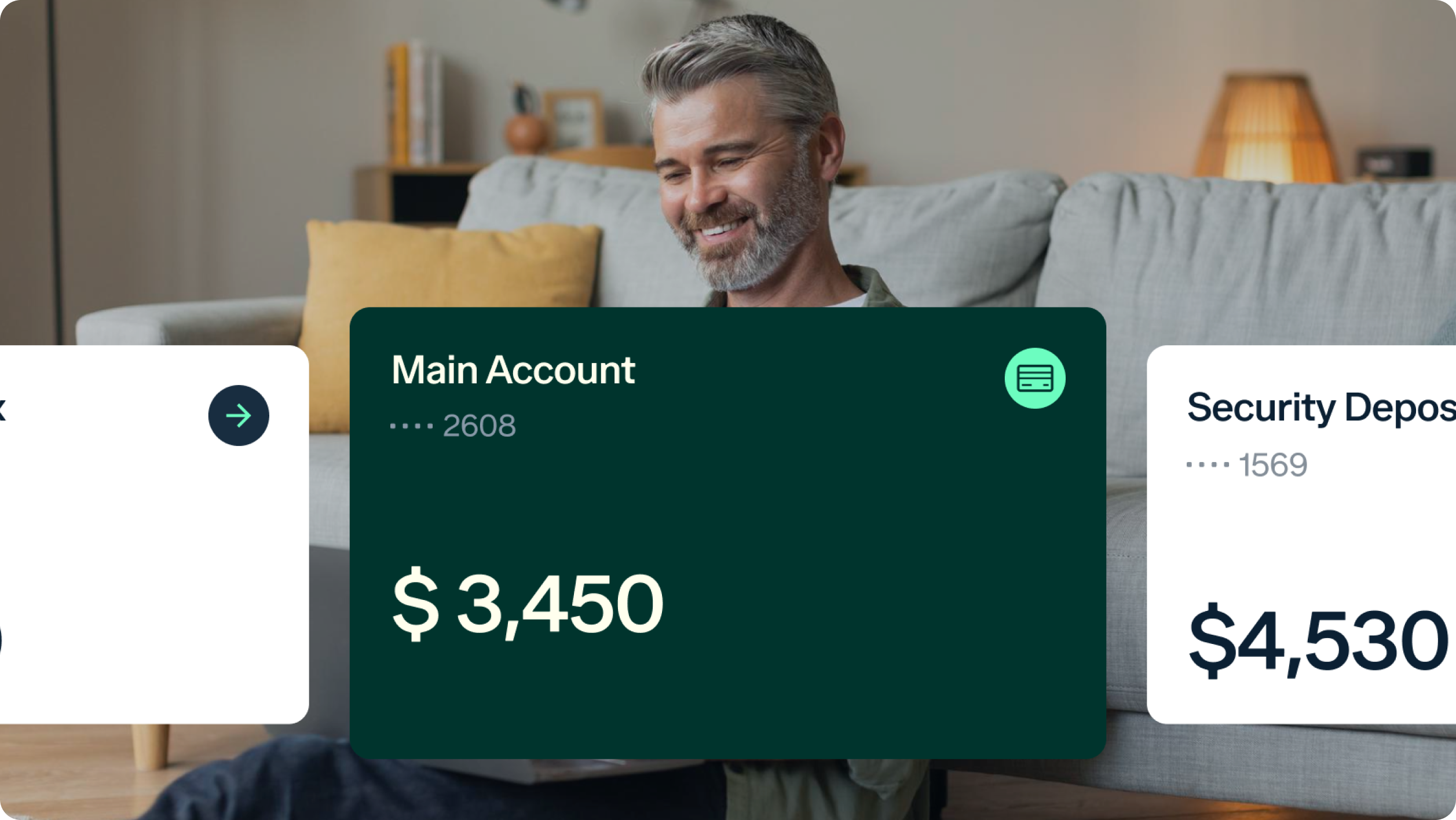 A man sits on the floor in a living room, leaning on a sofa with yellow pillows, using a laptop. Overlaid are digital card graphics showing account balances: Main Account with $3,450 and Security Deposit with $4,530.