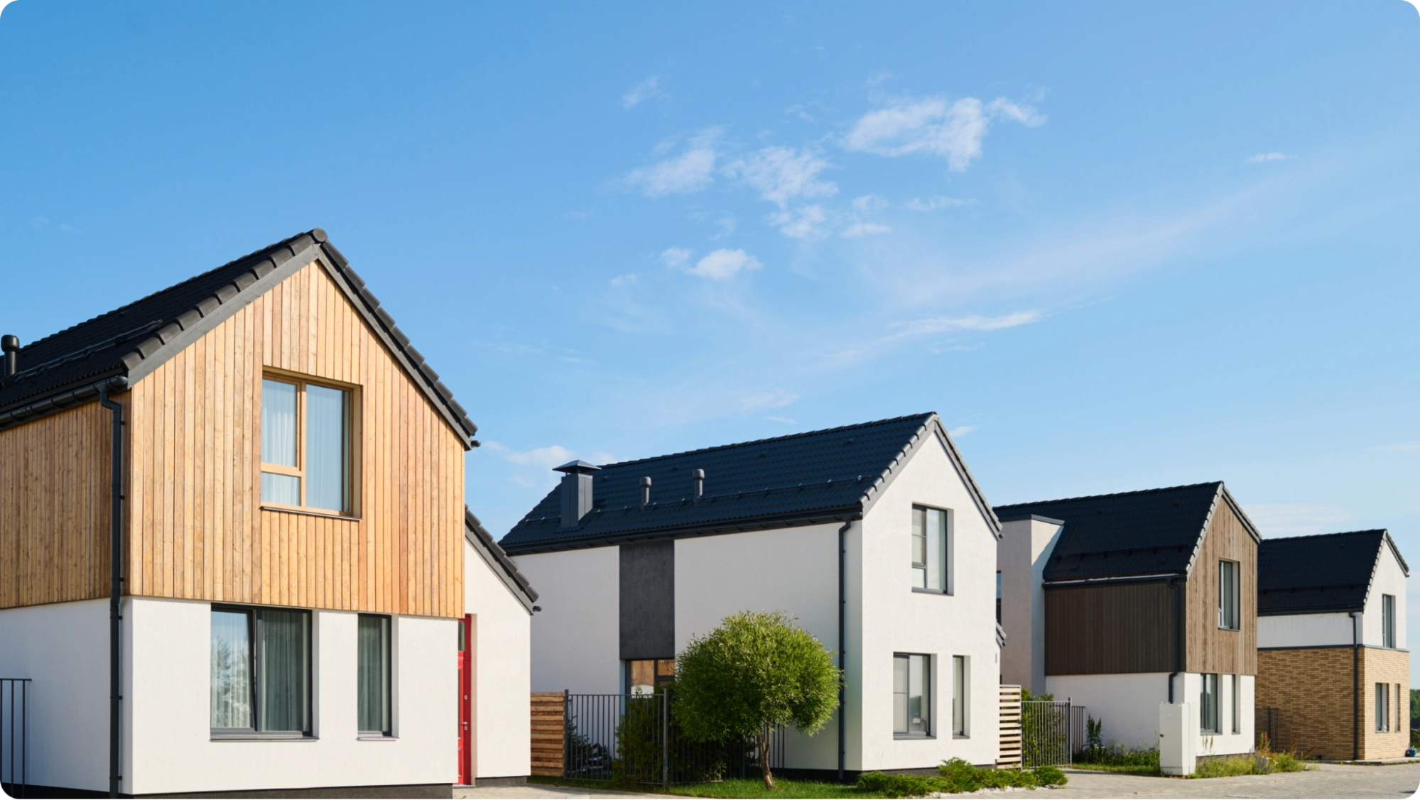 A row of modern houses with gable roofs under a clear blue sky. The front house features a combination of wood and white siding, while adjacent houses have white exteriors with dark roofs. A small tree and grass are visible in the foreground.