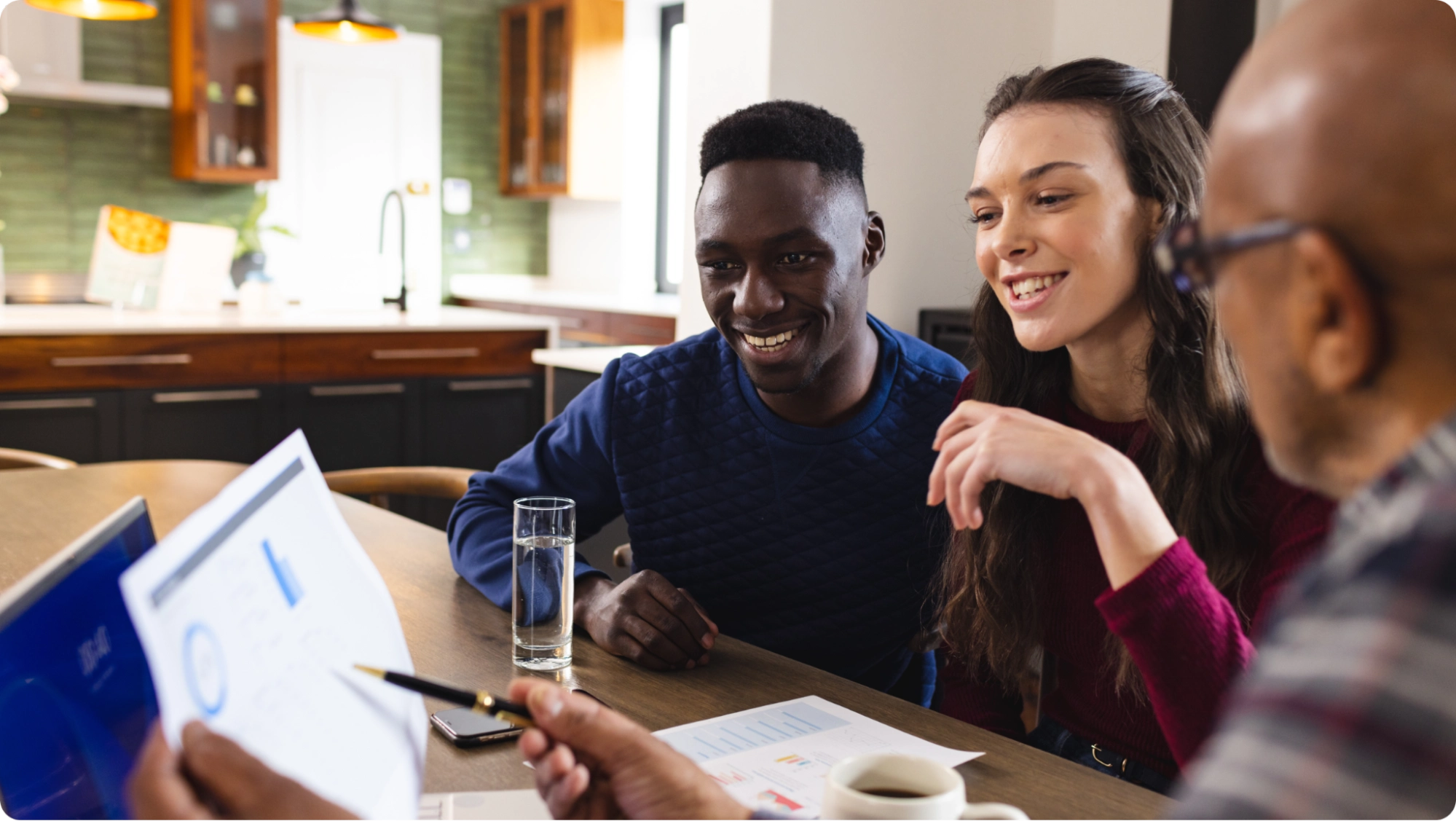 A group of people sit around a table discussing documents in a cozy kitchen setting. Two people are smiling and looking at charts, while someone points to the papers. There are glasses of water and a cup of coffee on the table.