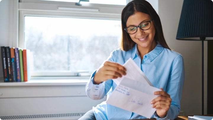 A woman with long hair and glasses smiles while opening a letter. She is seated by a window with a bookshelf in the background. She is wearing a light blue shirt and appears happy.