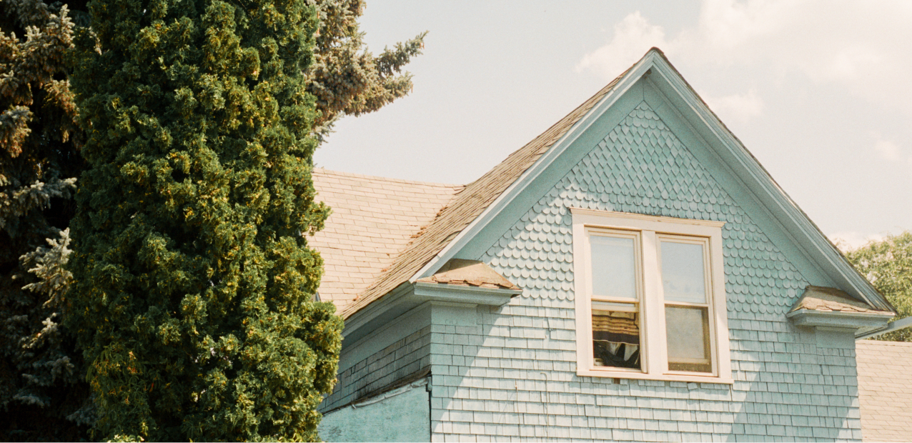 A close-up of the upper part of a light blue wooden house with a steep roof and a window. The exterior features shingle siding. A tall evergreen tree stands to the left of the house, and part of the blue sky with a few clouds is visible in the background.