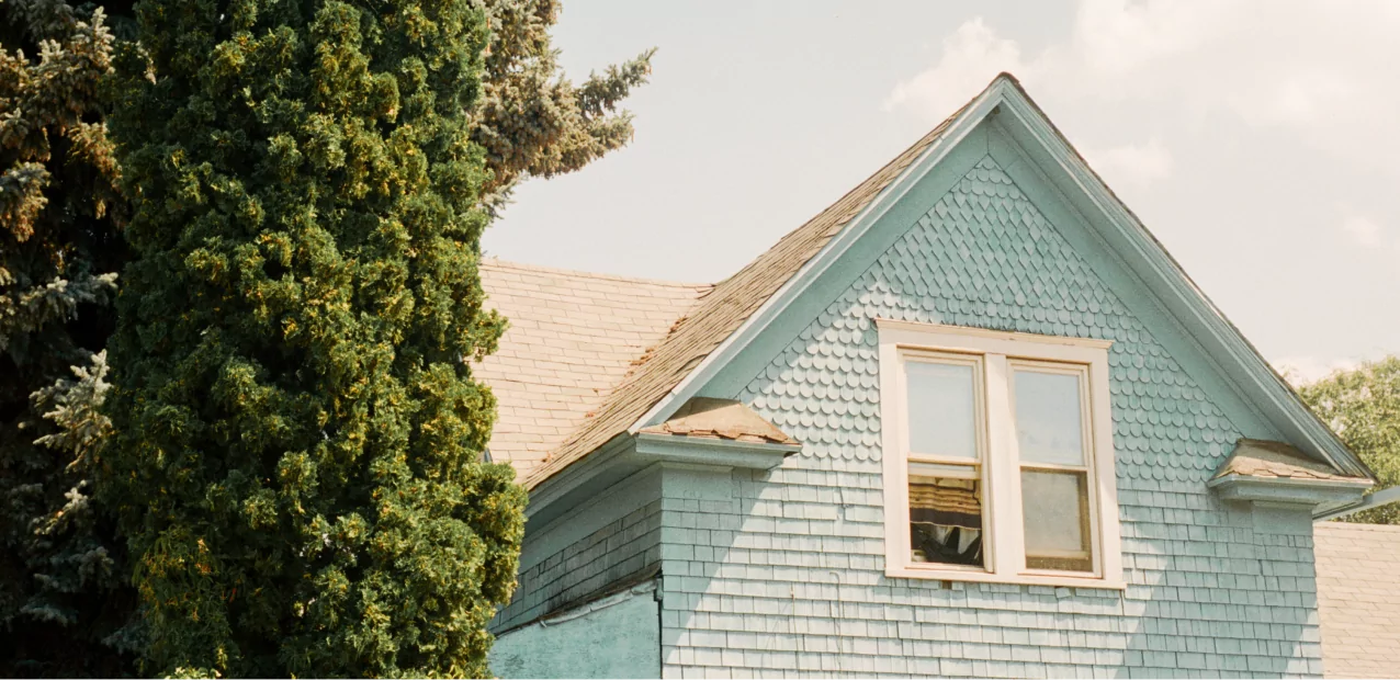 A close-up of the upper part of a light blue wooden house with a steep roof and a window. The exterior features shingle siding. A tall evergreen tree stands to the left of the house, and part of the blue sky with a few clouds is visible in the background.