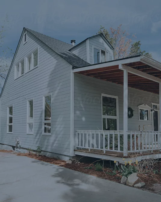 A light blue wooden house with white trim and a steep, dark roof sits in daylight. The home has small bushes in front, a raised porch with white railings, and several windows. The sky is clear with some leafy tree branches visible.