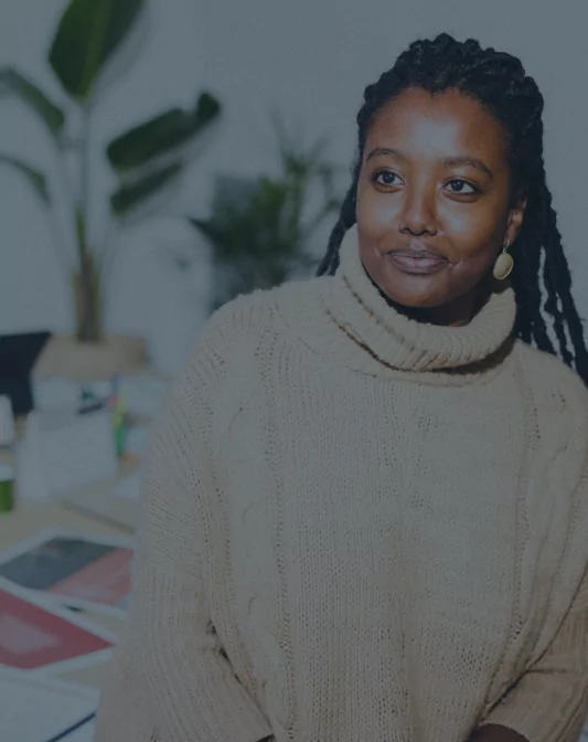 A person with long braided hair and gold earrings sits in an office space, wearing a beige knit sweater. The background includes plants, a desk with papers, and office supplies. The person has a calm, contemplative expression.