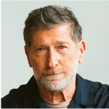 A middle-aged man with short, grey hair and a beard, wearing a navy blue shirt, looks directly at the camera against a plain light background.