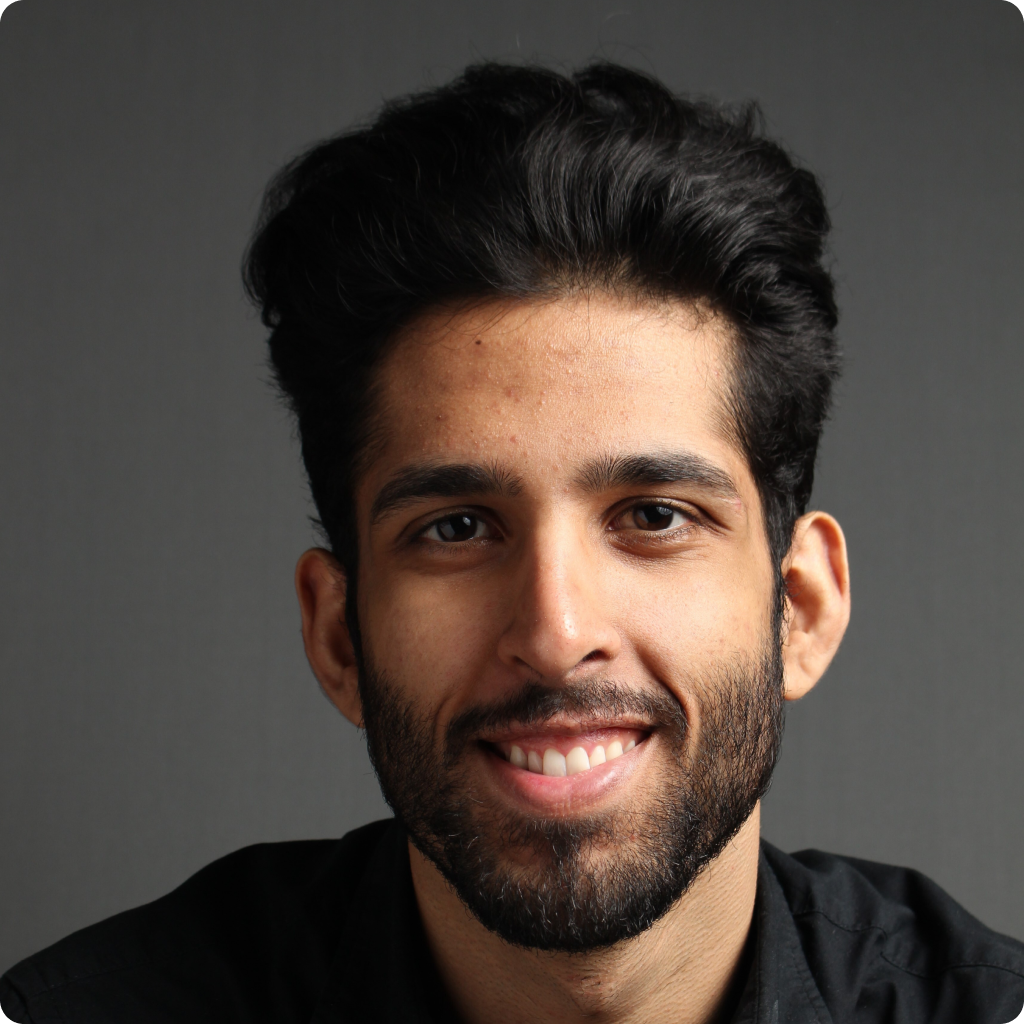 A person with dark hair and a beard smiles while wearing a black shirt, set against a gray background.