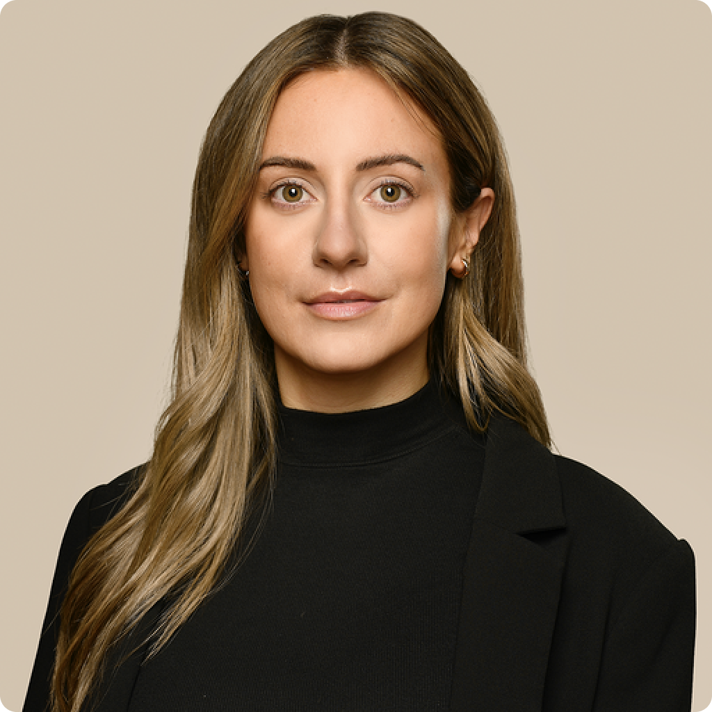 A woman with long blonde hair wearing a black turtleneck and blazer stands against a beige background. She looks directly at the camera with a neutral expression.