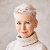 A woman with short, spiky white hair is smiling softly. She is wearing a cream-colored turtleneck sweater and small stud earrings. The background is a neutral, light-colored wall.