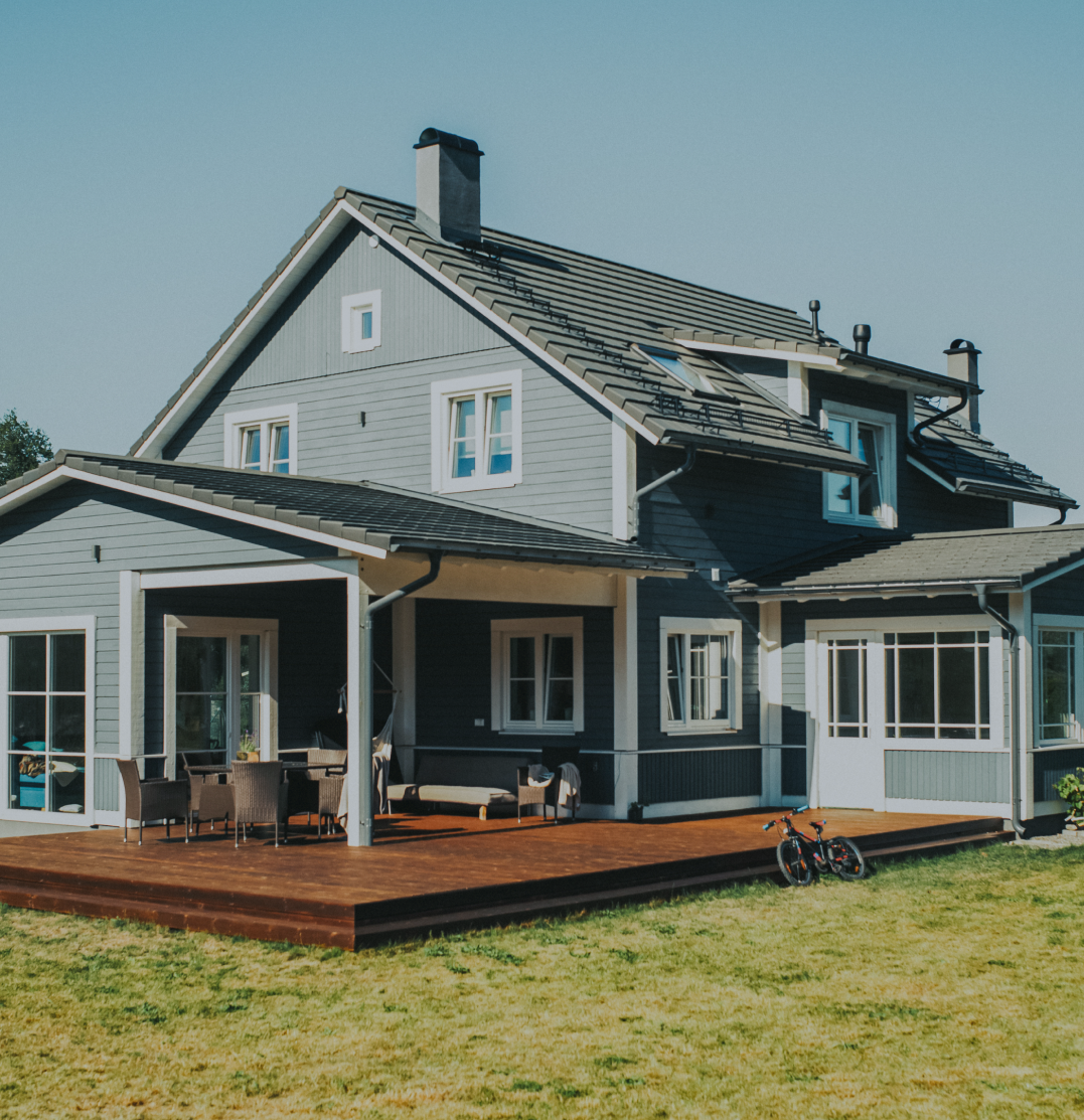 A two-story house painted in blue and white with a dark gray roof. The house features large windows and a spacious wooden deck with outdoor furniture. A bicycle is placed on the grass near the deck. The sky is clear, indicating a fair weather day.