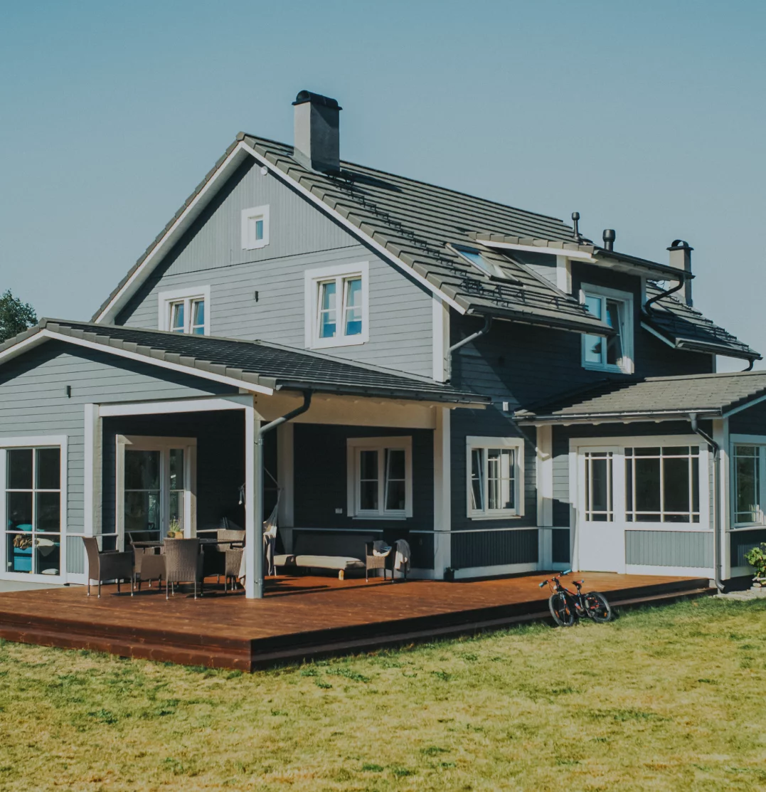 A two-story house painted in blue and white with a dark gray roof. The house features large windows and a spacious wooden deck with outdoor furniture. A bicycle is placed on the grass near the deck. The sky is clear, indicating a fair weather day.