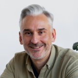A middle-aged man with short gray hair and a beard smiles warmly. He is wearing a light green button-up shirt and sits in front of a white background, possibly indoors. A partial view of a green plant can be seen on the right side of the image.
