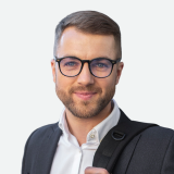 A man with short hair, a beard, and glasses is wearing a white-collared shirt under a blazer and carrying a shoulder bag. He is smiling and looking directly at the camera against a plain light background.