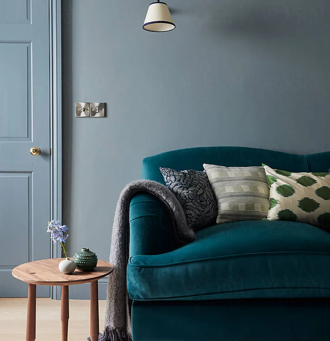 A cozy living room features a teal sofa adorned with patterned cushions and a woolen throw. A small wooden side table holds a vase with blue flowers and a decorative object. The backdrop includes a light blue door and a wall-mounted lamp.