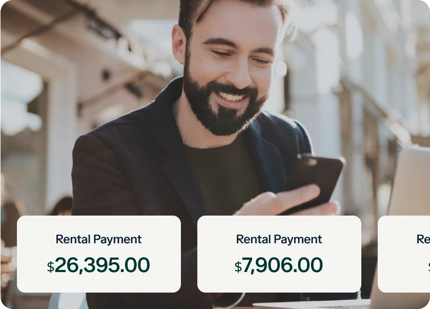 A bearded man is smiling and looking at his phone while holding a credit card. He is seated at an outdoor table with a laptop. In the foreground, there are two boxes with text: "Rental Payment $26,395.00" and "Rental Payment $7,906.00".