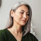 A woman with long, straight gray hair is looking down, wearing a green sweater. The background is blurred, focusing attention on her face, which appears thoughtful and calm.