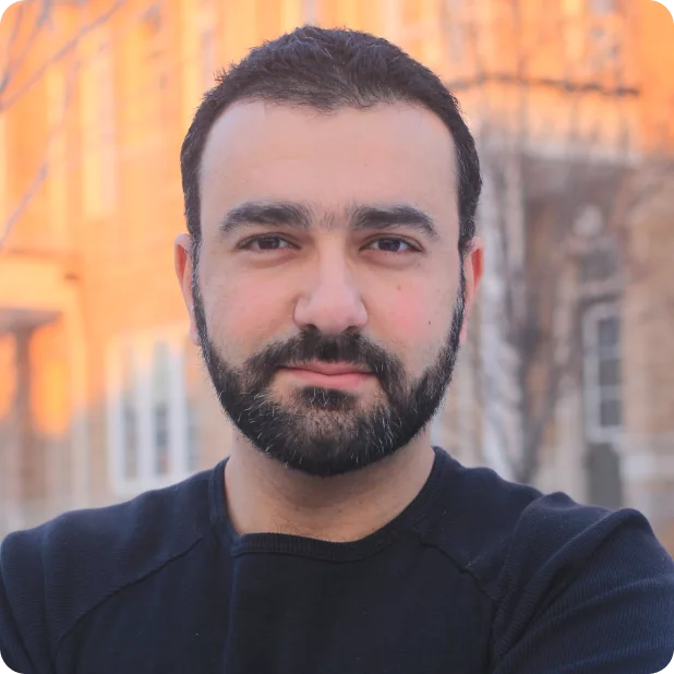 A person with a beard and short hair standing outside, facing the camera. They have a neutral expression and are wearing a black sweater. The background shows a building with warm sunlight casting an orange glow.
