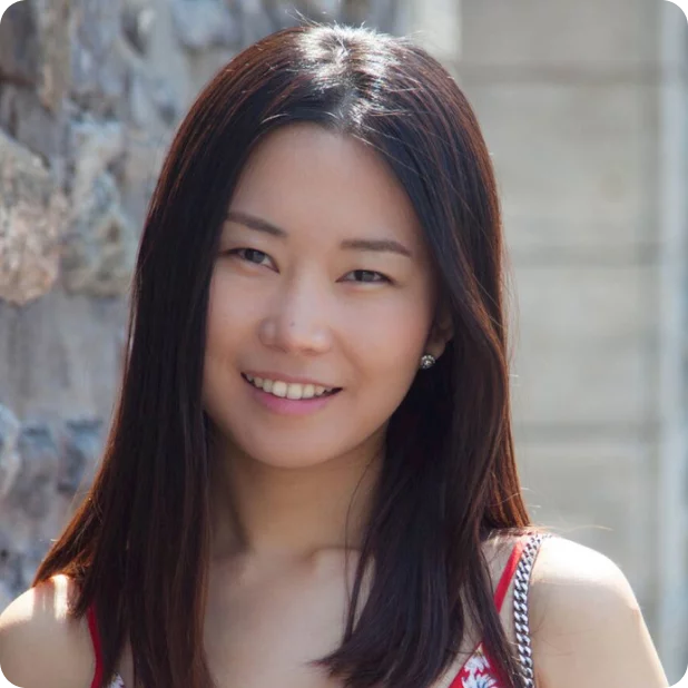 A woman with long, straight dark hair and wearing a sleeveless top smiles at the camera. She stands against a background featuring a stone wall and a building with a large window.