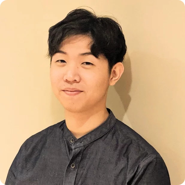 A young person with short black hair is smiling gently at the camera. They are wearing a dark button-up shirt and standing against a beige background. The lighting is soft and even.