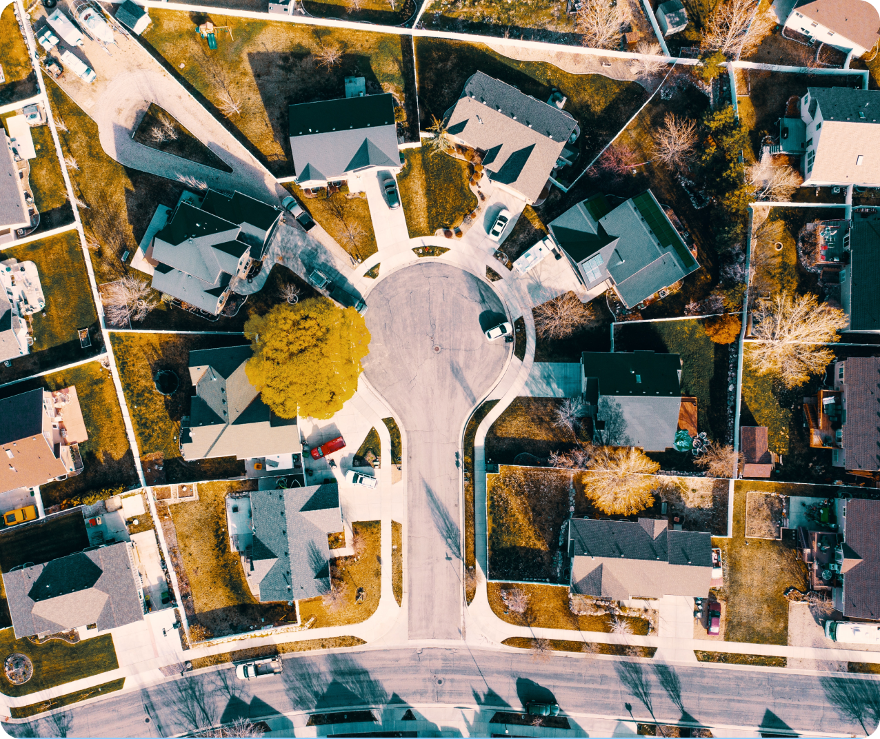 Aerial view of a suburban neighborhood with houses arranged around a cul-de-sac. Several homes have backyards and driveways. Trees, grass, and cars are visible, casting shadows in the sunlight. The streets are clean and well-defined.