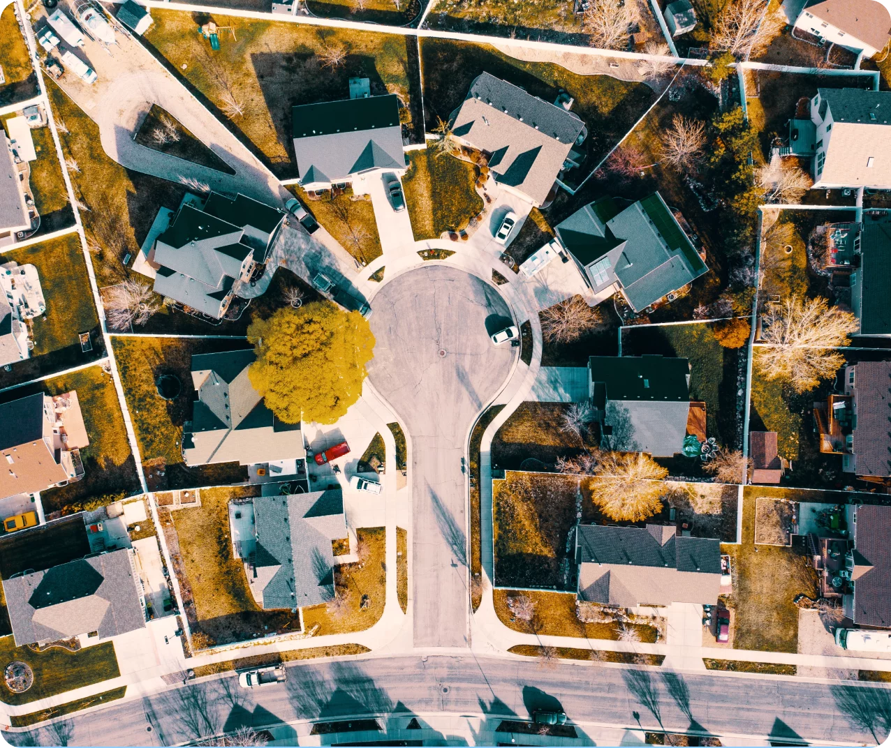 Aerial view of a suburban neighborhood with houses arranged around a cul-de-sac. Several homes have backyards and driveways. Trees, grass, and cars are visible, casting shadows in the sunlight. The streets are clean and well-defined.