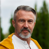 A man with short gray hair and a neatly trimmed beard is looking directly at the camera. He is wearing a white shirt and a yellow jacket. The background is slightly blurred, featuring green foliage.