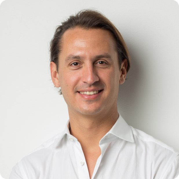 A person with medium-length hair is smiling and looking at the camera. They are wearing a white button-up shirt and standing against a plain light-colored background.