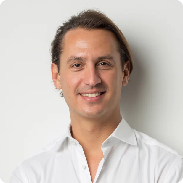 A person with medium-length hair is smiling and looking at the camera. They are wearing a white button-up shirt and standing against a plain light-colored background.