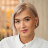 A person with short, platinum blonde hair and light skin is looking directly at the camera. They are wearing a white shirt and small earrings. The background is softly blurred, with warm tones and hints of yellow light.