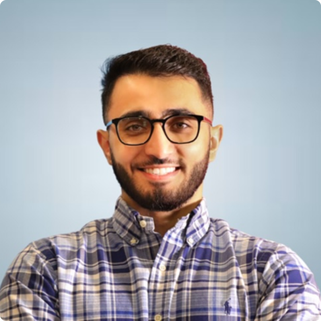 A smiling man with short dark hair and a beard, wearing glasses and a blue plaid shirt, stands against a light blue background.