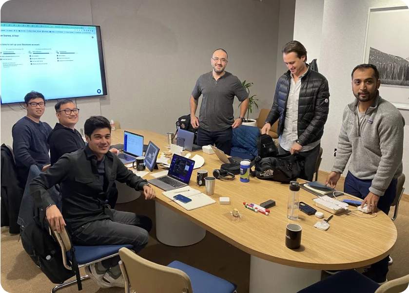 A group of six people are seated and standing around a table in a meeting room. Laptops, phones, coffee mugs, and notebooks are scattered on the table. A large screen on the wall displays a document. Everyone appears to be engaged and smiling.