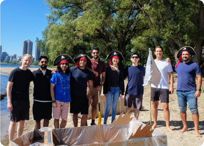 A group of nine people, some wearing pirate hats, stand on a sandy beach. They are posing for a photo next to a homemade cardboard boat. Trees are in the background, and tall buildings are visible in the distance. Everyone is smiling and the weather appears sunny.