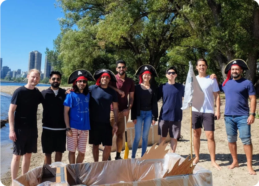 A group of nine people, some wearing pirate hats, stand on a sandy beach. They are posing for a photo next to a homemade cardboard boat. Trees are in the background, and tall buildings are visible in the distance. Everyone is smiling and the weather appears sunny.