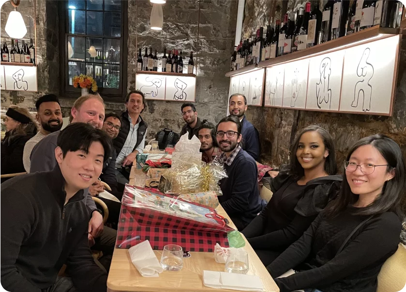 A group of eleven people sits around a long table in a cozy restaurant, smiling at the camera. Gift-wrapped presents are on the table. The walls are adorned with wine bottles and minimalist framed artwork. The setting is warm and inviting.