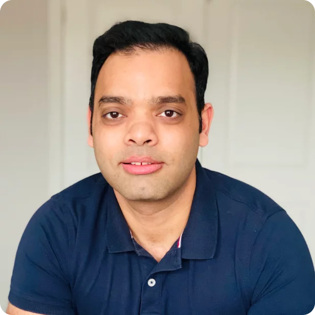 A person with short dark hair and a slight smile is wearing a dark blue polo shirt. They are posed against a blurred indoor background with a white door.