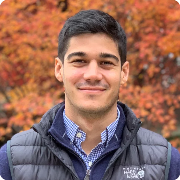 A person smiles at the camera, standing outdoors with vibrant autumn foliage in the background. They have short dark hair and are wearing a dark quilted vest over a blue checkered shirt with the words "Mountain Hard Wear" on the vest.