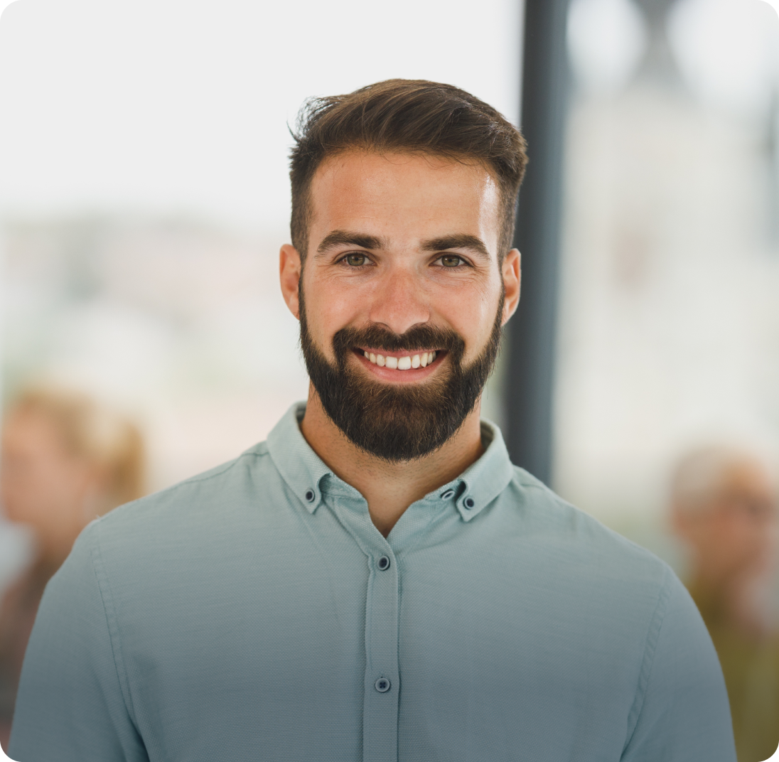 A man with a beard and short dark hair smiles at the camera. He is wearing a light teal button-up shirt. The background is blurred, showing other people and a hint of a building, suggesting an indoor setting.