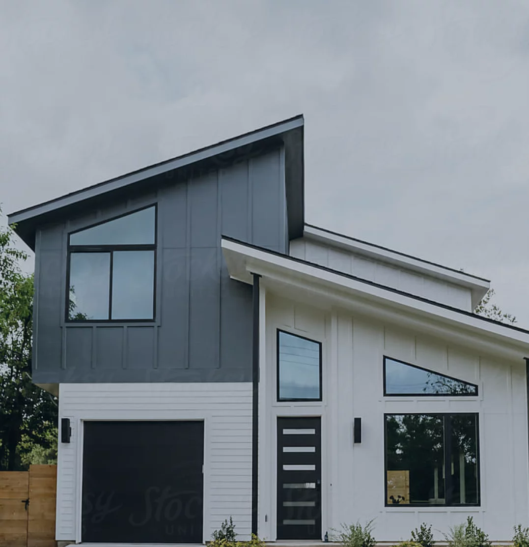 Modern two-story house with a combination of dark and light gray siding, large windows, and a sloped roof. The upper portion of the facade features vertical paneling, and the entrance has a stylish black door with multiple horizontal glass inserts.