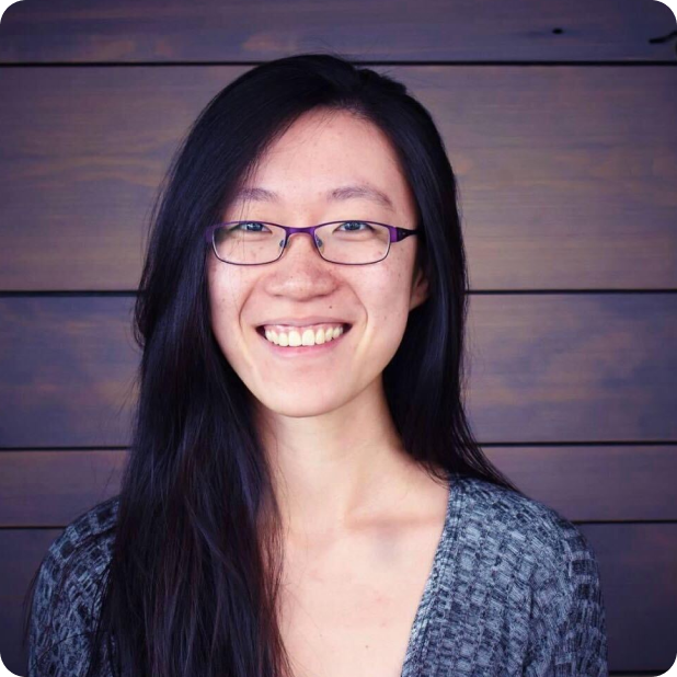 A person with long black hair and glasses is smiling. They are wearing a gray patterned shirt and standing in front of a wooden plank background.
