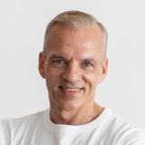 A middle-aged man with short, light hair smiles at the camera. He is wearing a plain white T-shirt, and the background is a simple, light gray color.