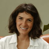 A woman with wavy dark hair smiles warmly at the camera. She is wearing a white collared shirt and is seated indoors, with a green plant leaf visible in the background.