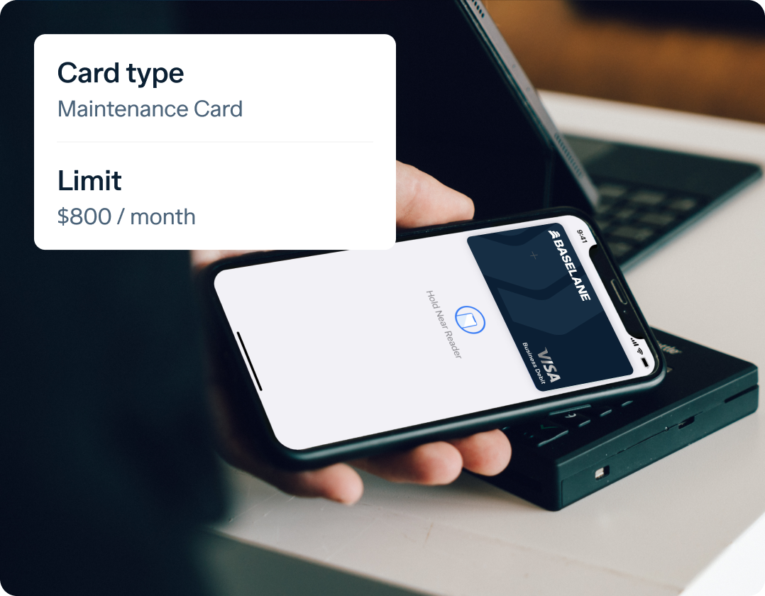 A person holding a smartphone displaying a virtual card with the label "MAINTENANCE VISA." Below the card, “Card type: Maintenance Card” and “Limit: $800 / month” are shown. In the background, a laptop and a docking station are visible.