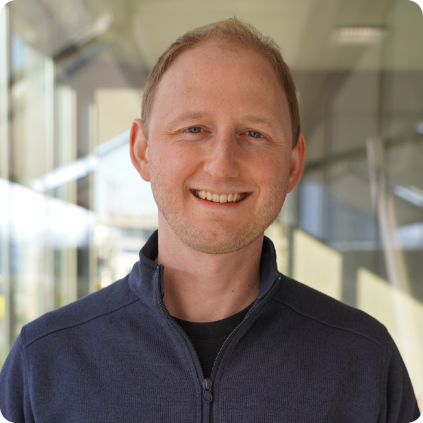 A man with short light hair is smiling while wearing a dark blue zip-up jacket. He stands indoors with large glass windows and bright natural light in the background.