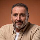 A middle-aged man with short dark hair, a trimmed beard, and a mustache is posed against a smooth, burnt orange background. He wears a beige button-down shirt layered over a white t-shirt and has a relaxed, composed expression.