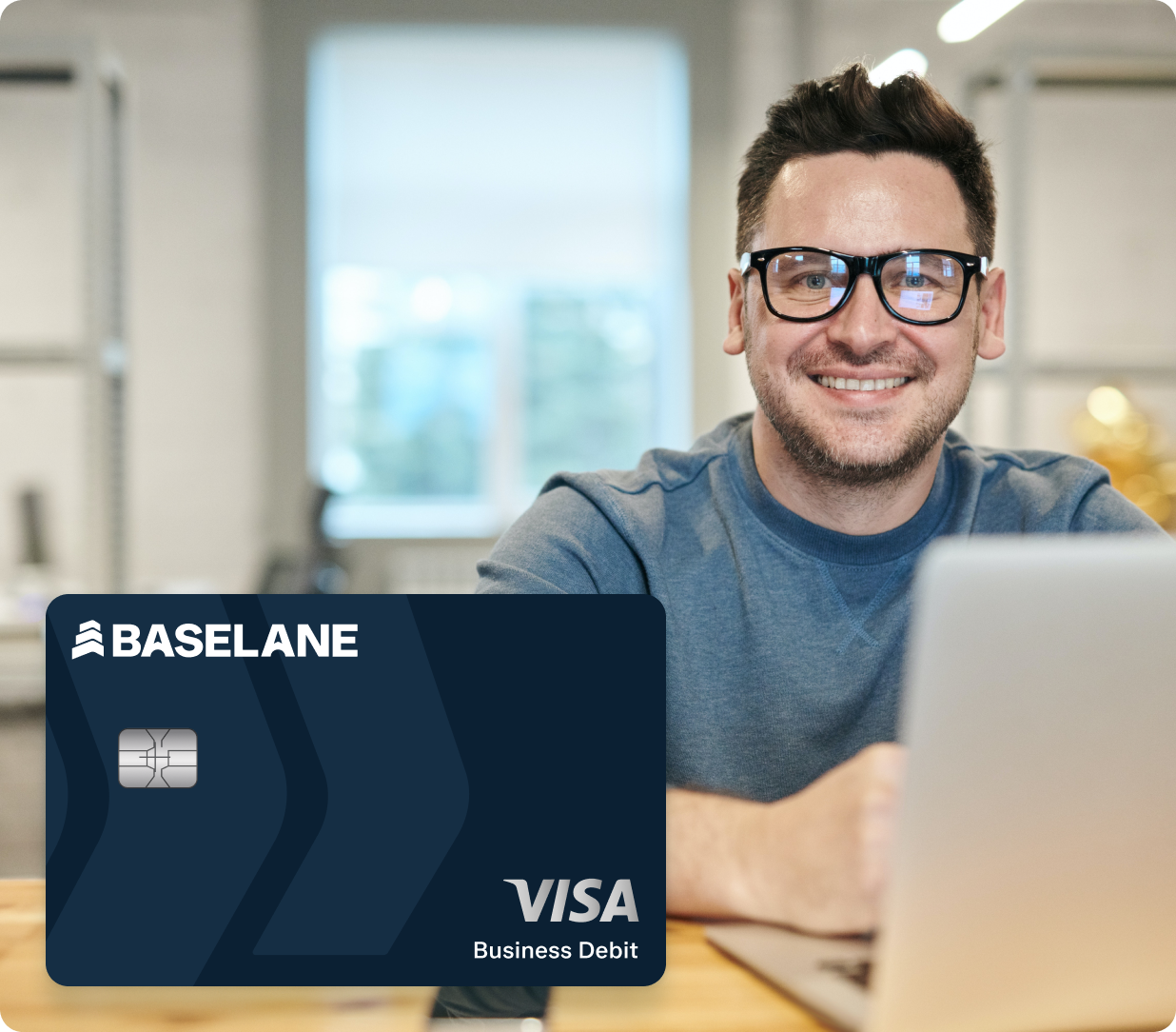 A person wearing glasses and a grey shirt is smiling while working on a laptop in a modern office setting. In the foreground, there is a large image of a Baseline Visa Business Debit card.