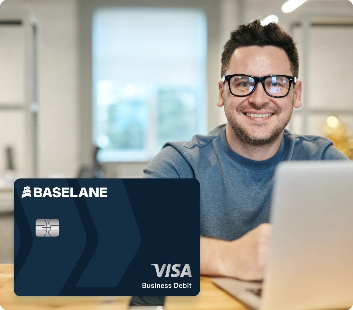 A person wearing glasses and a grey shirt is smiling while working on a laptop in a modern office setting. In the foreground, there is a large image of a Baseline Visa Business Debit card.