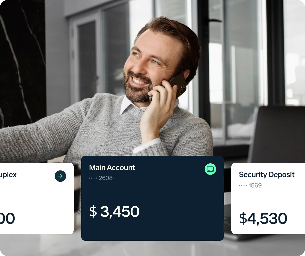 A man with a beard is smiling while talking on his phone in a modern office. He is wearing a gray sweater with a white collared shirt underneath. The screen also displays account information with amounts: $3,450 for Main Account and $4,530 for Security Deposit.