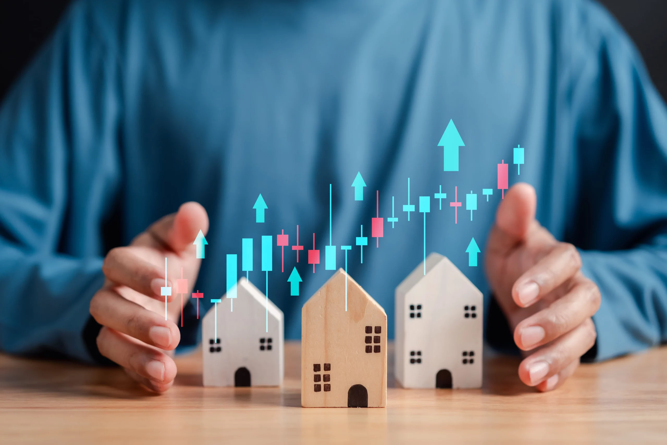 A person in a blue shirt with hands hovering over miniature wooden houses sits at a table. Overlaid on the image are colorful stock market graphs and arrows pointing upwards, symbolizing growth in the real estate market.